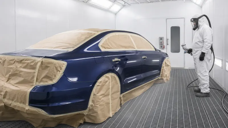 A masked navy sedan inside a white spray booth, with a painter in a full protective suit holding a spray gun